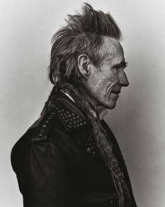 Black and white image of Peter Beaumont, facing to the right. He has spiked grey hair, wrinkles, and is wearing a studded leather jacket with a zip up cardigan beneath. 