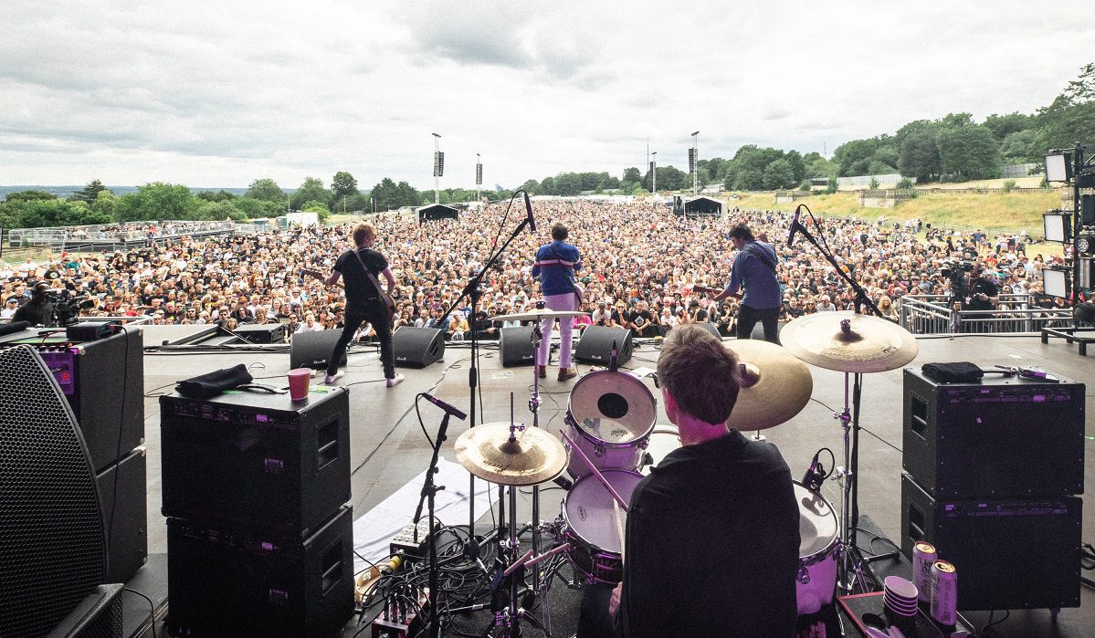 Image shows shot from back of the stage out across the crowds with Buzzcocks playing at the front, facing away from the photographer and towards the crowd