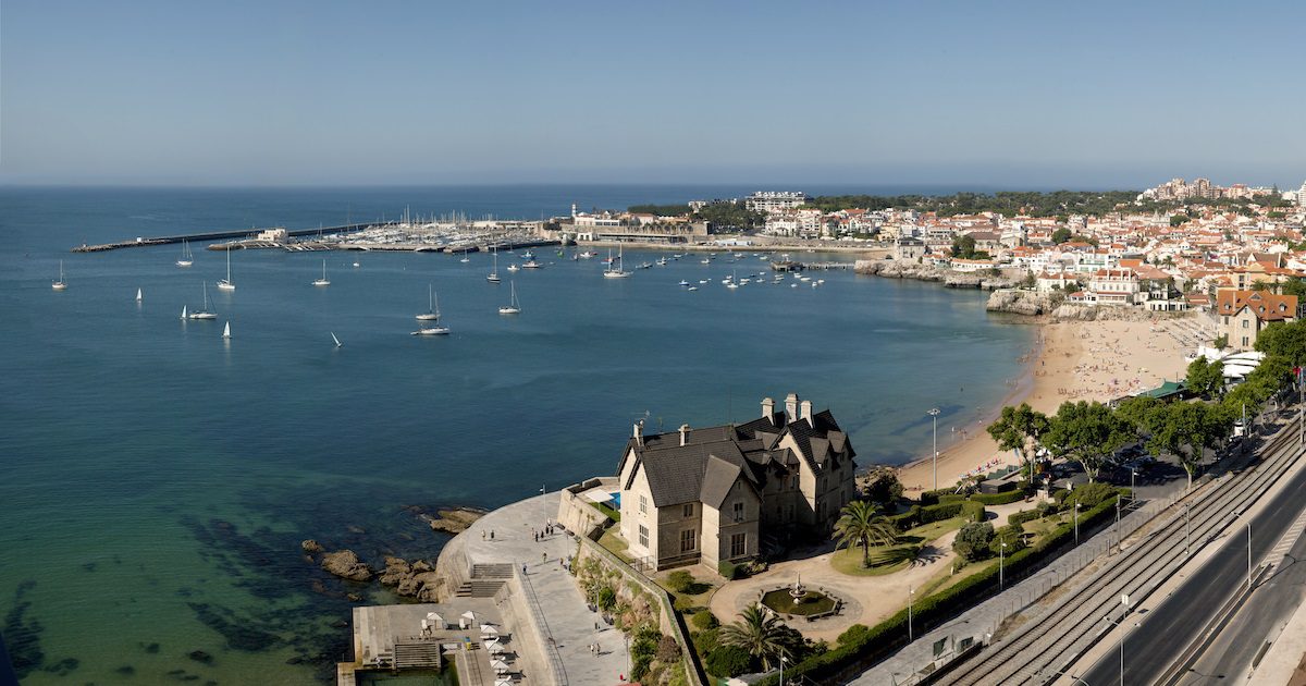 Beautiful blue cove with costal town building gathered around. A few boats float in the harbour. Taking a holiday alone to Cascais