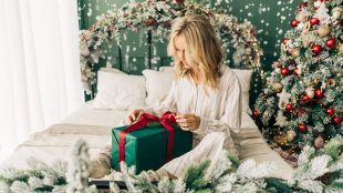 Woman in pyjamas smiling and untying ribbon on christmas gift box while sitting on comfortable decorated bed. Christmas alone article on Silver Magazine www.silvermagazine.co.uk