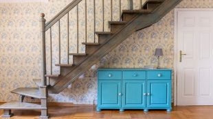A light blue three door cabinet standing under gray staircase against a wall with flower wallpaper in a hall interior. Convert cupboard under the stairs - Silver Magazine www.silvermagazine.co.uk