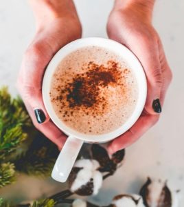 Two hands cupping a warm mug with cinnamon on top, and pine needles on the surface below.