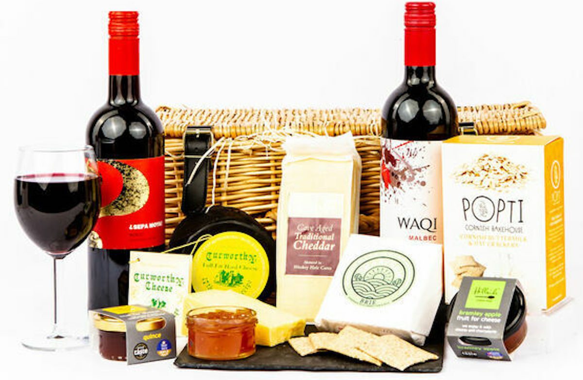 Hamper against a white background with bottles of wine and cheese displayed in front of it.