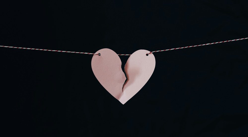 image shows pink paper heart on black background. Paper heart is torn, representing heartbreak