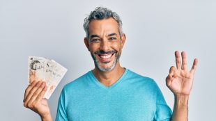 Middle age grey-haired man holding united kingdom pounds doing ok sign with fingers, smiling friendly gesturing excellent symbol. eBay side hustle now without sellers fees - article on Silver Magazine https://silvermagazine.co.uk/side-hustle-ebay-drops-sellers-fees