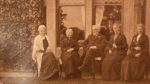 A victorian photograph showing five people in historical clothing sat on chairs outside a home.