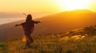 Image shows woman happy and carefree feeling awestruck by a beautiful sunset, arms raised
