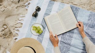A book is open on a beach towel surrounded by sand. someone is holding the pages open and there are sunglasses to the left of the book on the towel