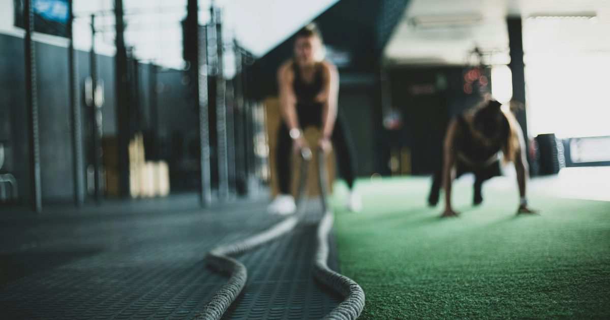 A woman is at the gym in dark clothing crouching down holding long work out ropes. The floor is dark green and there is a grey matt.