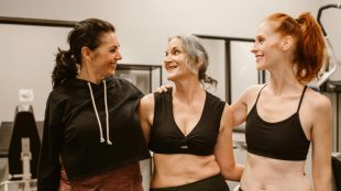 Three women stand side by side in gym clothing with their arms around each other. They are in a gym setting.