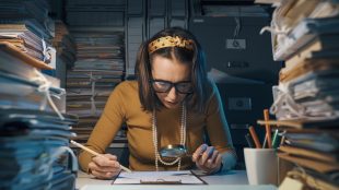 Handling a loved ones estate advice Silver Magazine. Woman sitting at office desk and checking a contract with a magnifier, she is surrounded by piles of paperwork