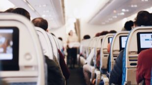 Image of the aisle of an aeroplane, with people seated and a blurred person walking down the aisle.