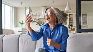 Happy mature 60s aged woman reacting to mobile phone message lottery winning big sum of money. Feeling great happiness of receiving gift on sofa at home. How would you feel if you won the lottery Silver Magazine www.silvermagazine.co.uk