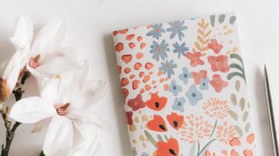 Floral print notebook on a table with flowers next to it and a pen on the right hand side. Learn the benefits of journalling on Silver