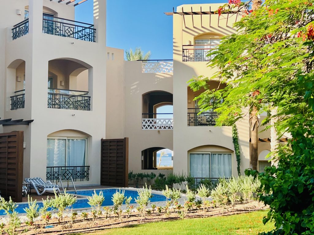 The image shows the hotel. It is cream and has multiple large windows and balconeys. There is a pool underneath and a single deck chair.