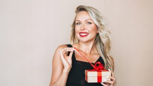 Portrait of gorgeous happy blonde young woman with bright makeup in evening dress with gift box in hands on beige background. Last minute Valentine gifts for her that she'll love - Silver Magazine www.silvermagazine.co.uk