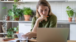 Sad contemplative woman examining bills on laptop. Overdue pay car finance, gas, electricity, etc financial hardship concept, with glass of water in home kitchen. Financial stress, debt issues, household budget finance struggles, overdue accounts, money problems. Missold car finance Silver Magazine www.silvermagazine.co.uk