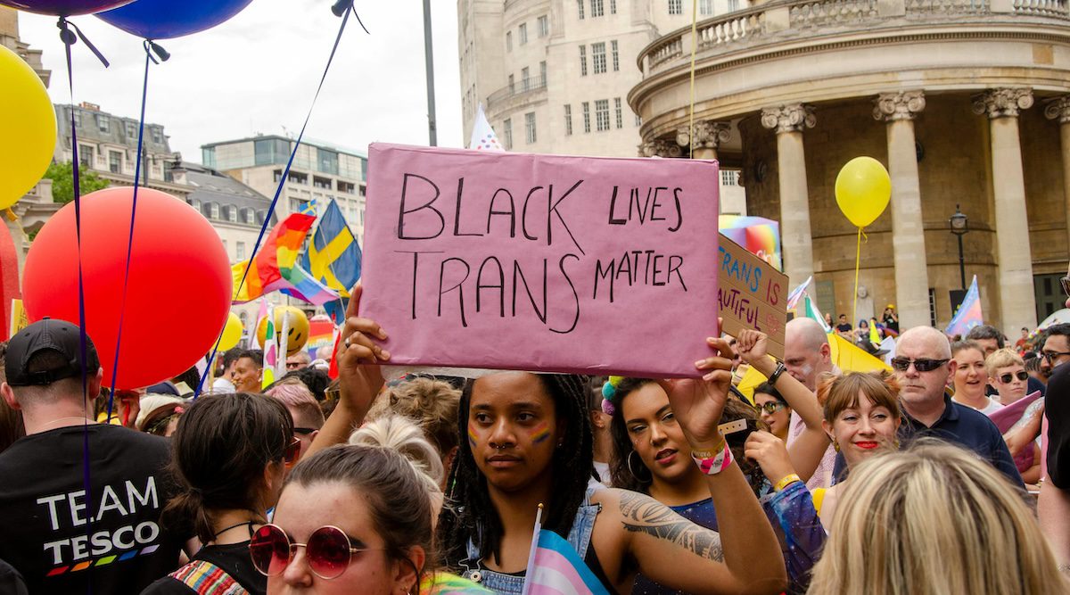 Person at a Pride crowd in Britain holds up a sign that reads 'Black Trans Lives Matters', a man in the back wears a shirt that says 'Team Tesco' with pride colours on it. It's Pride Month - exploring Pride in the UK, silvermagazine.co.uk