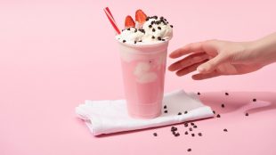 Partial view of female hand with disposable cup of milkshake with chocolate morsels and strawberry on napkins on pink.