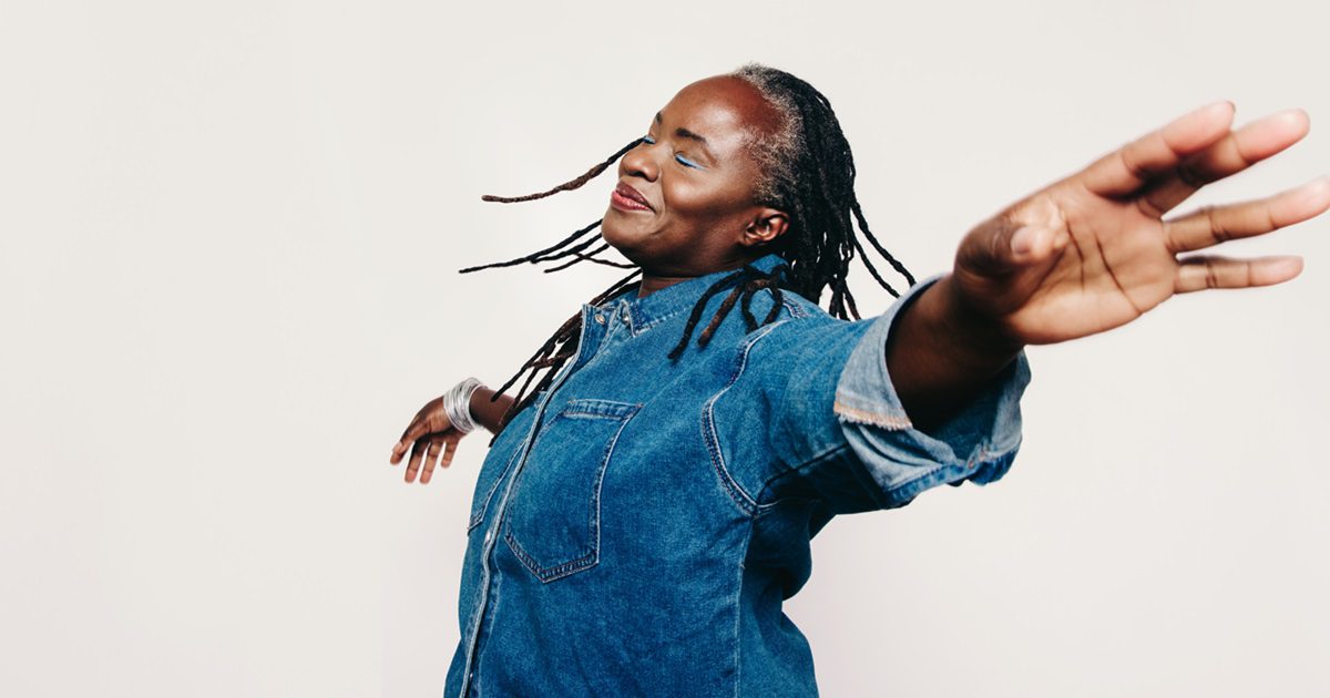 Mature happiness. Carefree woman with dreadlocks standing with her eyes closed and her arms outstretched. Cheerful middle-aged woman wearing a denim jacket and make-up in a studio.