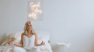 Woman sat in white pyjamas meditation on bed with 'Good Vibes Only' light up sign behind her