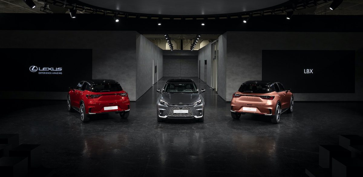 A red, grey and gold Lexus LBX under a spotlight in a car show room.