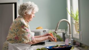 An elderly woman wearing an apron washing carrots under a sink tap in front of the window. Helping ageing parents nutrition tips on Silver