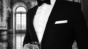 A close up view of a man in a black tuxedo holding a whiskey glass in a mansion. Why dressing up matters - Silver Magazine www.silvermagazine.co.uk