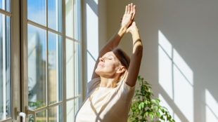 Older woman basking in the sunlight from her window, arms stretched up, eyes closed.