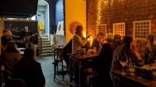 Image taken inside YADA Collective bar, where a group of people are sat around a dimly lit table with a performer behind them on their left.