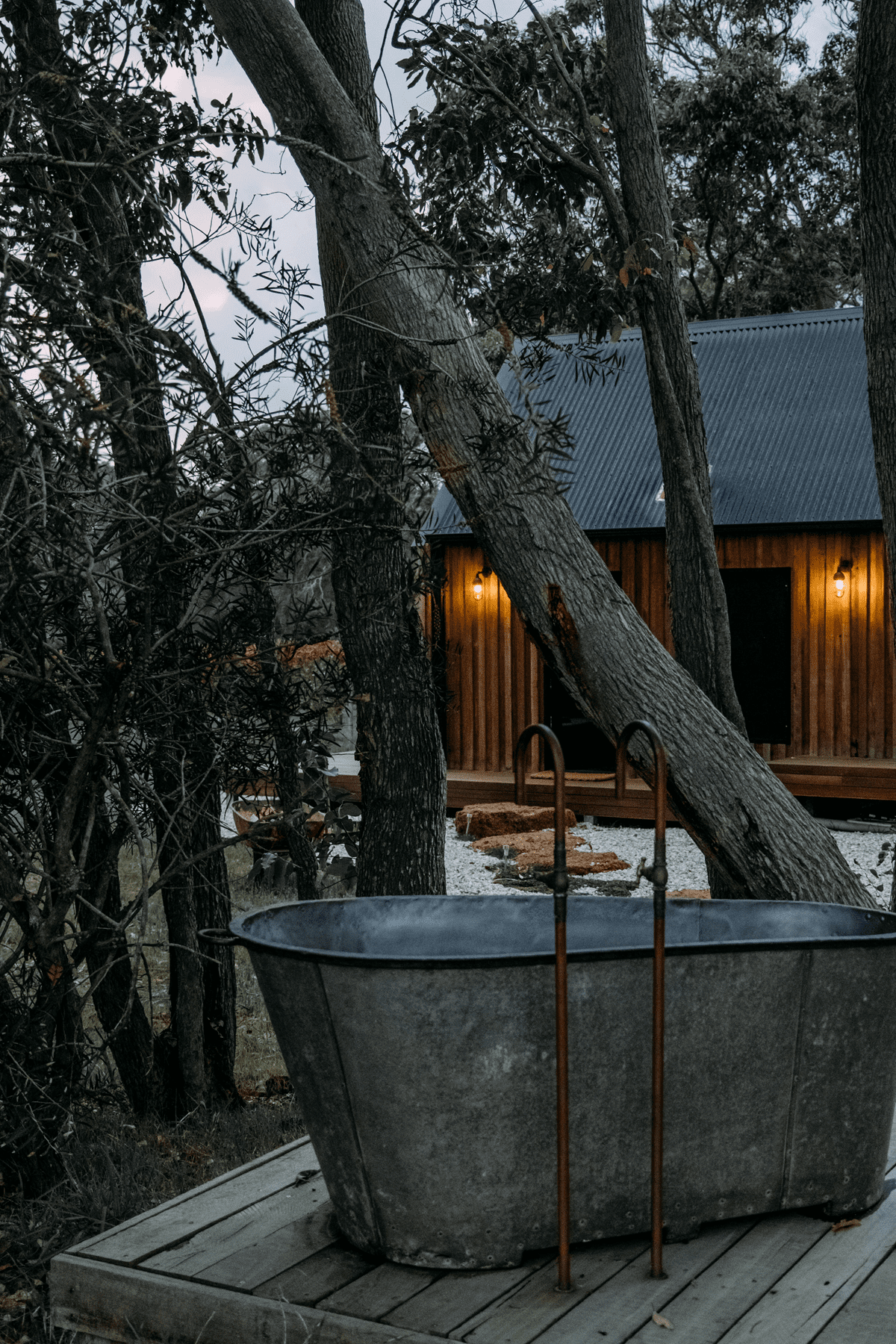 A wide shot of a rusty bathtub with a dimly lit cabin in the background, surrounded by forest trees.