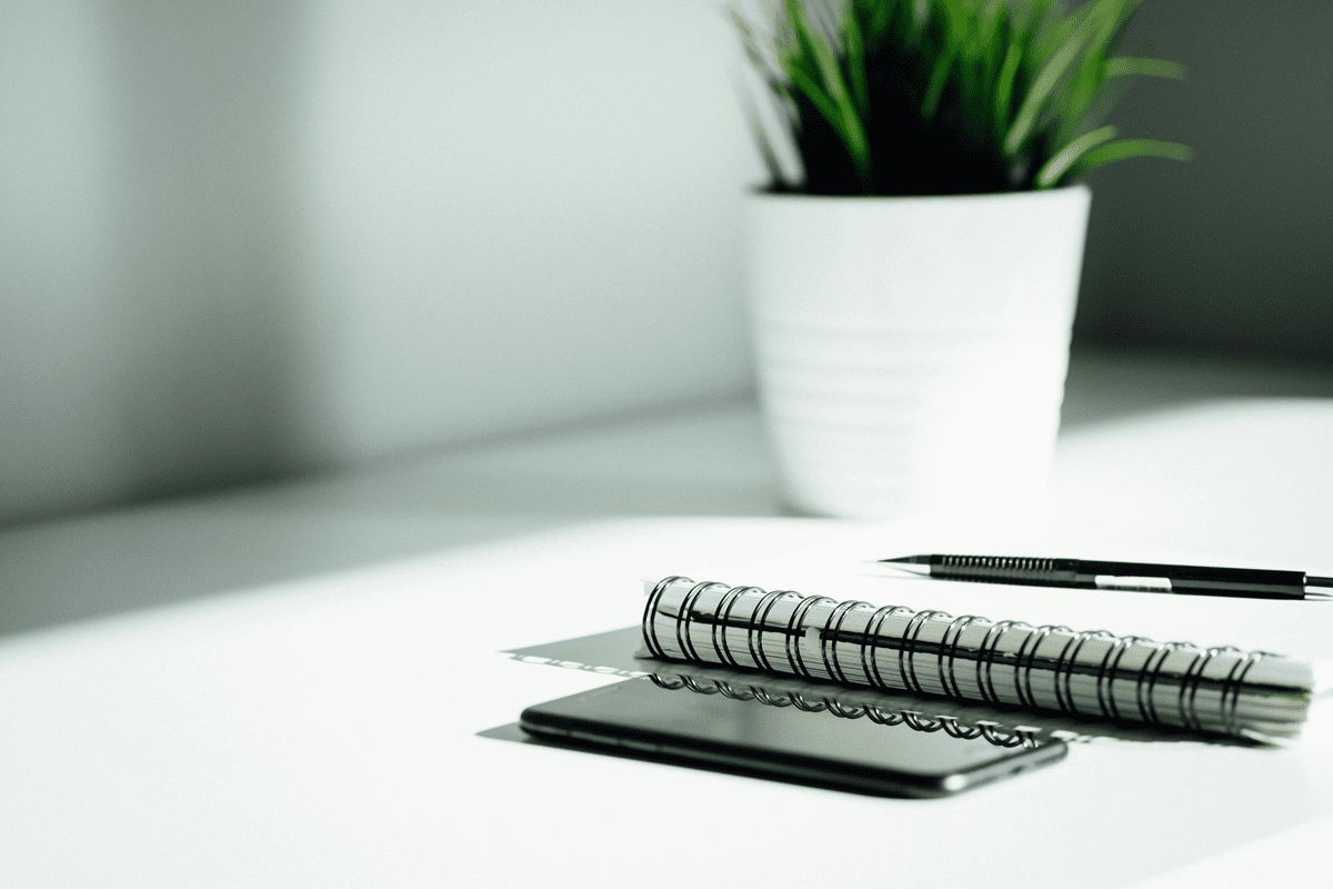 A close up of a white desk with with a notepad with a black pen on top. Next to the notepad there is a black mobile phone. in the background there is a green plant in a white verse. 